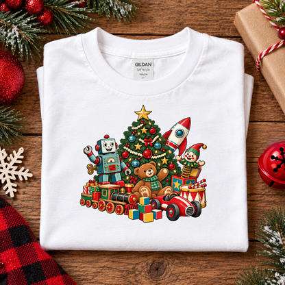 A folded white Gildan Softstyle T-shirt is displayed on a rustic wooden surface, decorated with Christmas-themed props.  The front of the shirt features a colorful graphic of a Christmas tree with toys, including a robot, a teddy bear, a rocket, and a toy car.