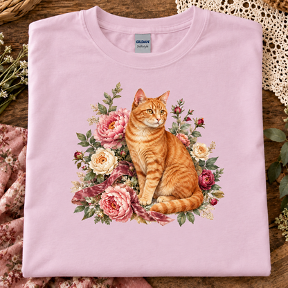A folded light pink Gildan t-shirt with a graphic print of an orange tabby cat surrounded by pink and cream roses, displayed on a rustic wooden surface with decorative lace and fabric.