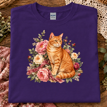 A folded purple Gildan t-shirt featuring a detailed graphic of an orange tabby cat surrounded by pink and cream roses, displayed on a rustic wooden surface with lace and floral props.