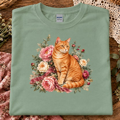 A folded sage green t-shirt with a detailed graphic of an orange tabby cat surrounded by pink and cream roses, displayed on a rustic wooden surface with decorative lace and flowers.
