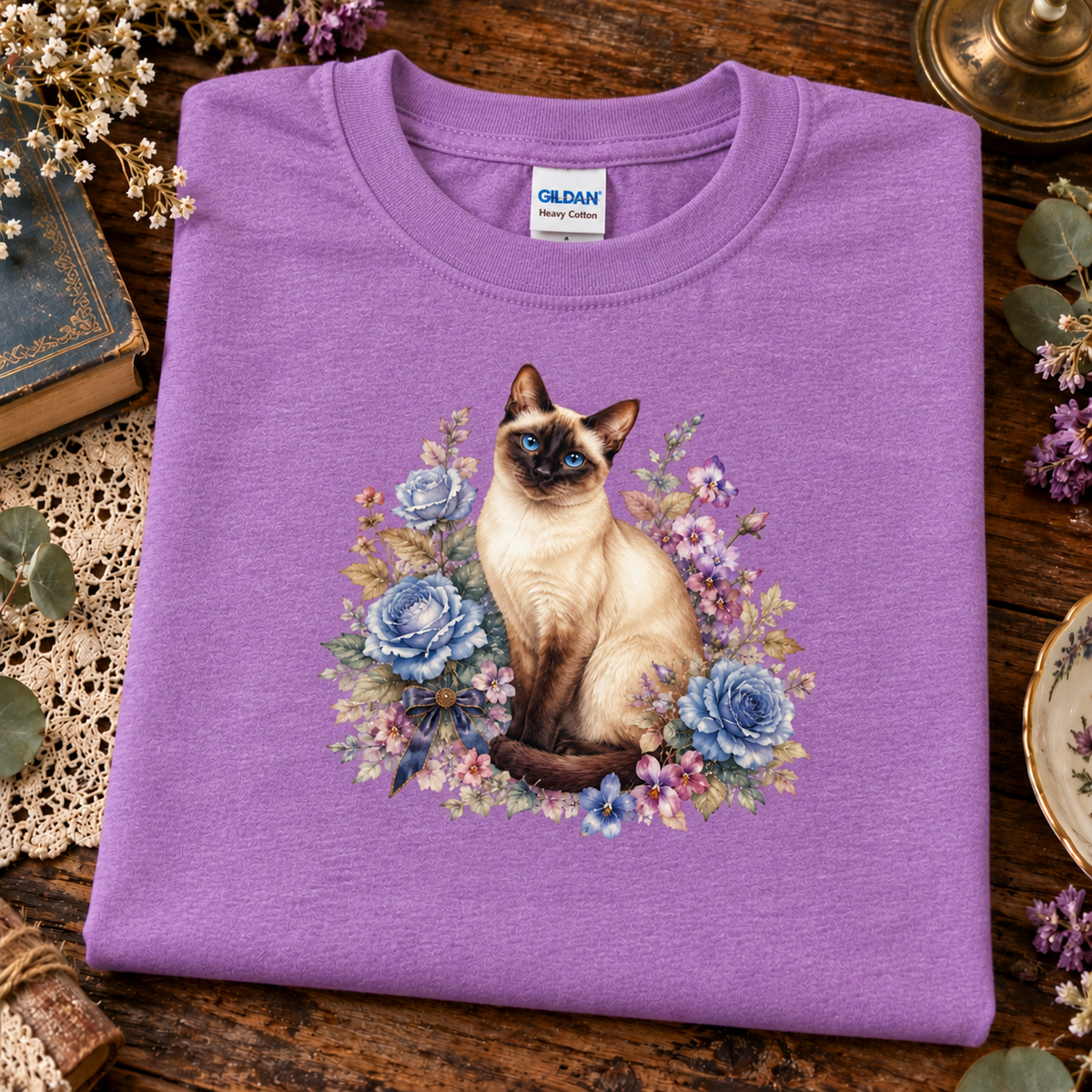 A folded heather radiant orchid crewneck sweatshirt with a graphic of a Siamese cat surrounded by blue and purple flowers, displayed on a rustic wooden table with decorative props.