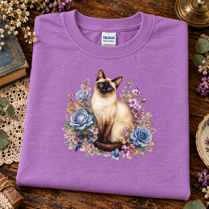 A folded heather radiant orchid crewneck sweatshirt with a graphic of a Siamese cat surrounded by blue and purple flowers, displayed on a rustic wooden table with decorative props.
