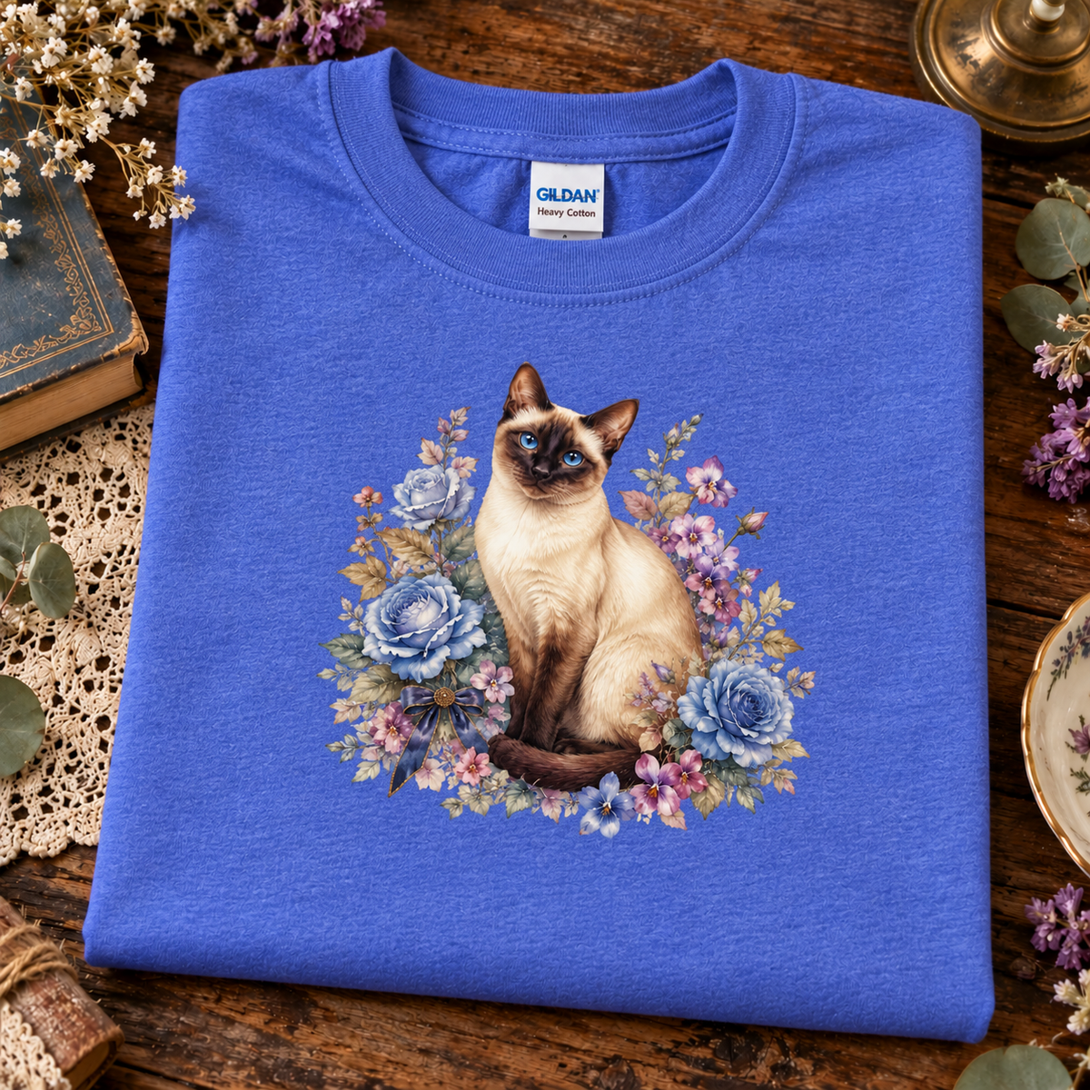A folded heather blue Gildan t-shirt with a graphic of a Siamese cat surrounded by blue and purple flowers, displayed on a rustic wooden table with decorative floral props.