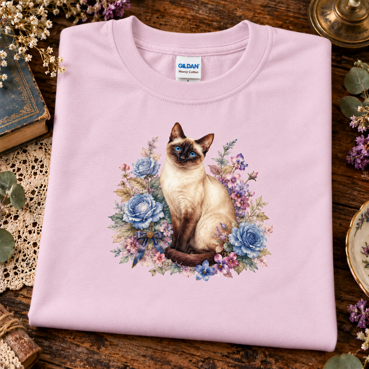 A folded light pink Gildan t-shirt with a graphic of a Siamese cat surrounded by blue and purple flowers, displayed on a rustic wooden table with vintage props.