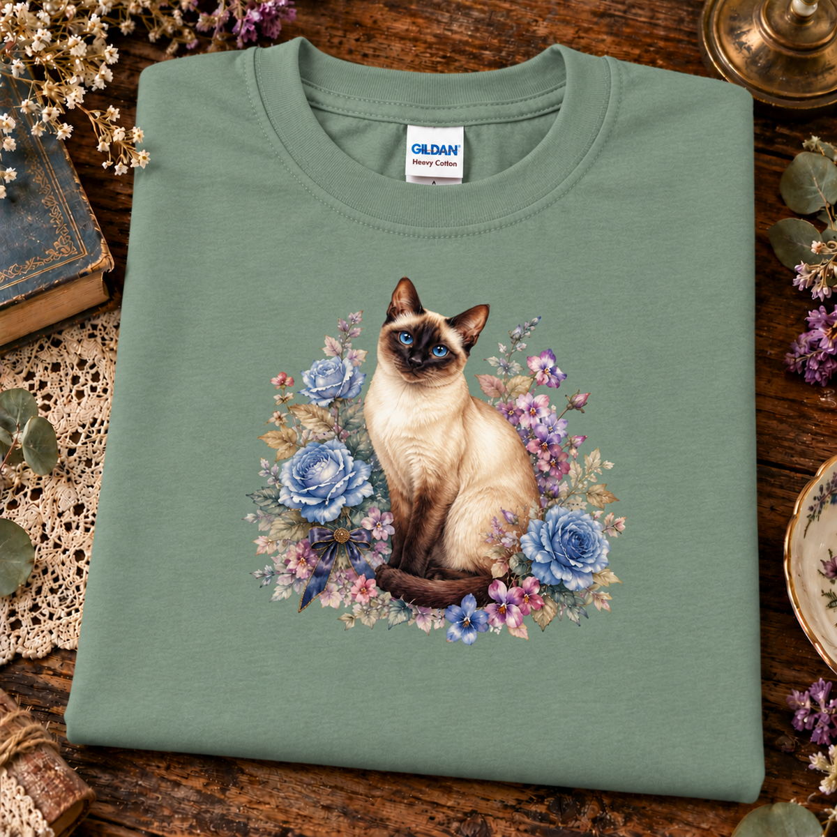 A folded sage green Gildan t-shirt featuring a detailed graphic of a Siamese cat surrounded by blue and purple flowers, displayed on a rustic wooden table with vintage props.