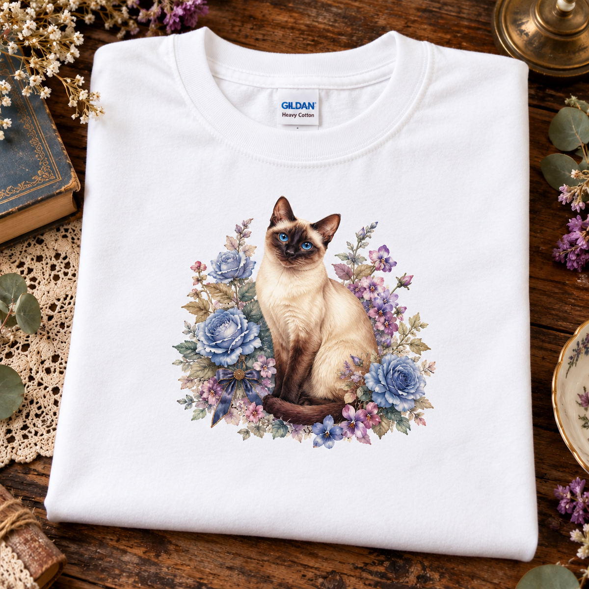 A folded white Gildan Heavy Cotton t-shirt featuring a printed graphic of a Siamese cat surrounded by blue and purple flowers, displayed on a rustic wooden table with decorative props.
