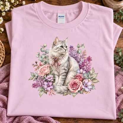 A folded light pink Gildan t-shirt with a detailed graphic of a gray tabby cat surrounded by pink and purple flowers, displayed on a wooden surface with a pink blanket.
