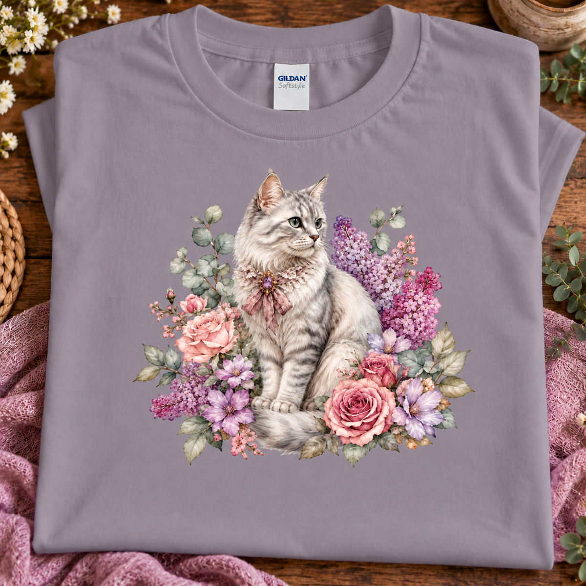 A folded paragon t-shirt with a detailed graphic of a fluffy gray cat surrounded by pink and purple flowers, displayed on a rustic wooden surface with a pink blanket and greenery.