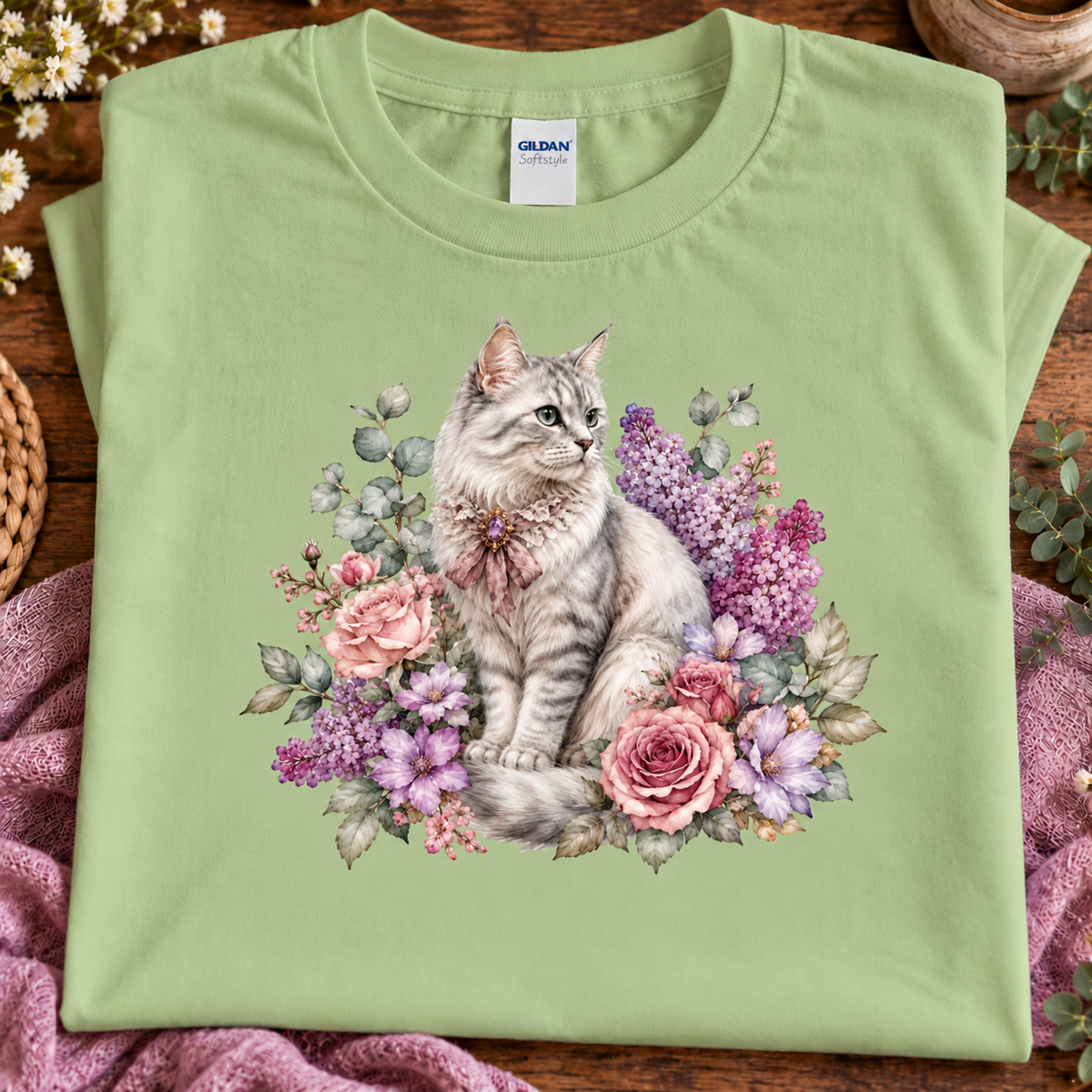 A folded pistachio green t-shirt with a detailed graphic of a fluffy gray tabby cat surrounded by pink and purple flowers, displayed on a rustic wooden surface with a pink blanket.