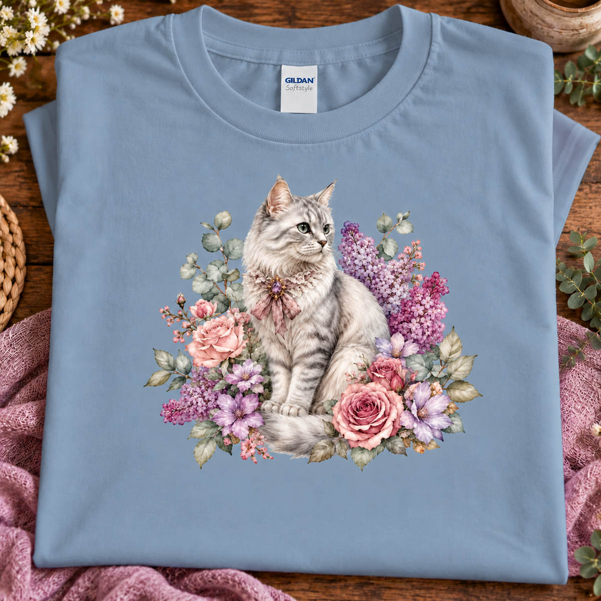 A folded stone blue Gildan t-shirt with a detailed graphic of a gray tabby cat surrounded by pink and purple flowers, displayed on a wooden surface with a pink blanket.