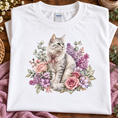 A folded white Gildan t-shirt with a detailed graphic of a gray tabby cat surrounded by pink and purple flowers, displayed on a wooden surface with a pink blanket.