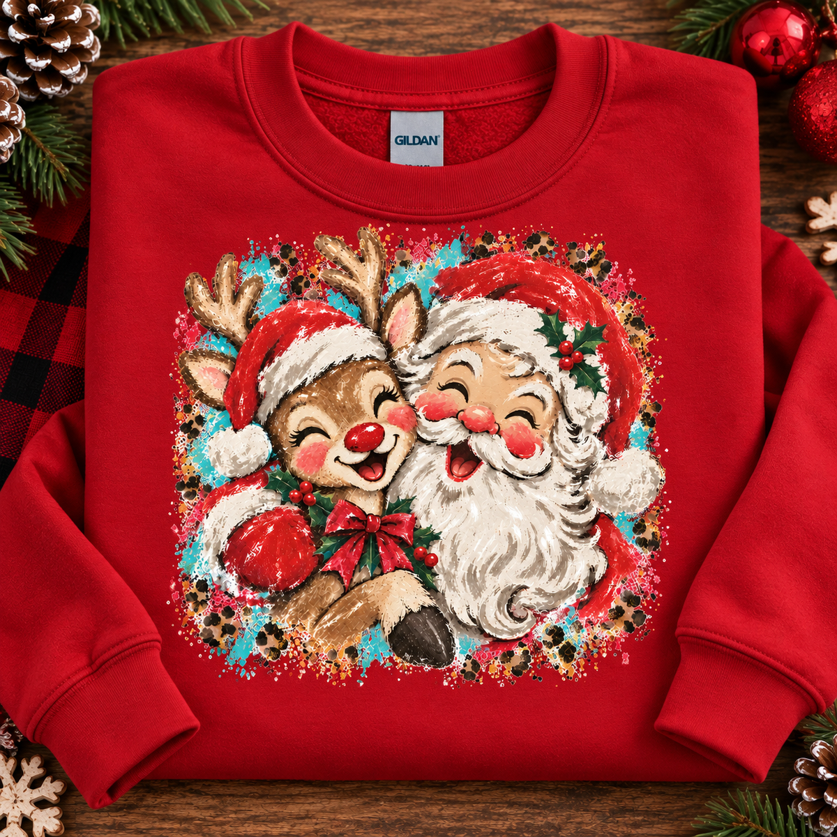 A red crewneck sweatshirt laid flat on a wooden surface, decorated with holiday greenery, pinecones, and ornaments. The front features a large graphic of a smiling reindeer and Santa Claus with red hats, white fur, and a festive multicolor background.