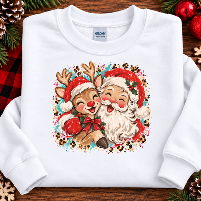 A white Gildan sweatshirt laid flat on a rustic wooden surface, decorated with pine branches, red ornaments, and a plaid blanket. The sweatshirt features a colorful graphic of a Santa Claus and a reindeer hugging, framed by a leopard-print border.
