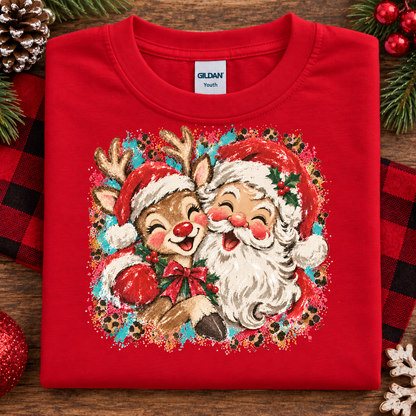 A folded red Gildan Youth t-shirt with a colorful Christmas graphic of a reindeer and Santa Claus, styled on a wooden surface with holiday decorations.