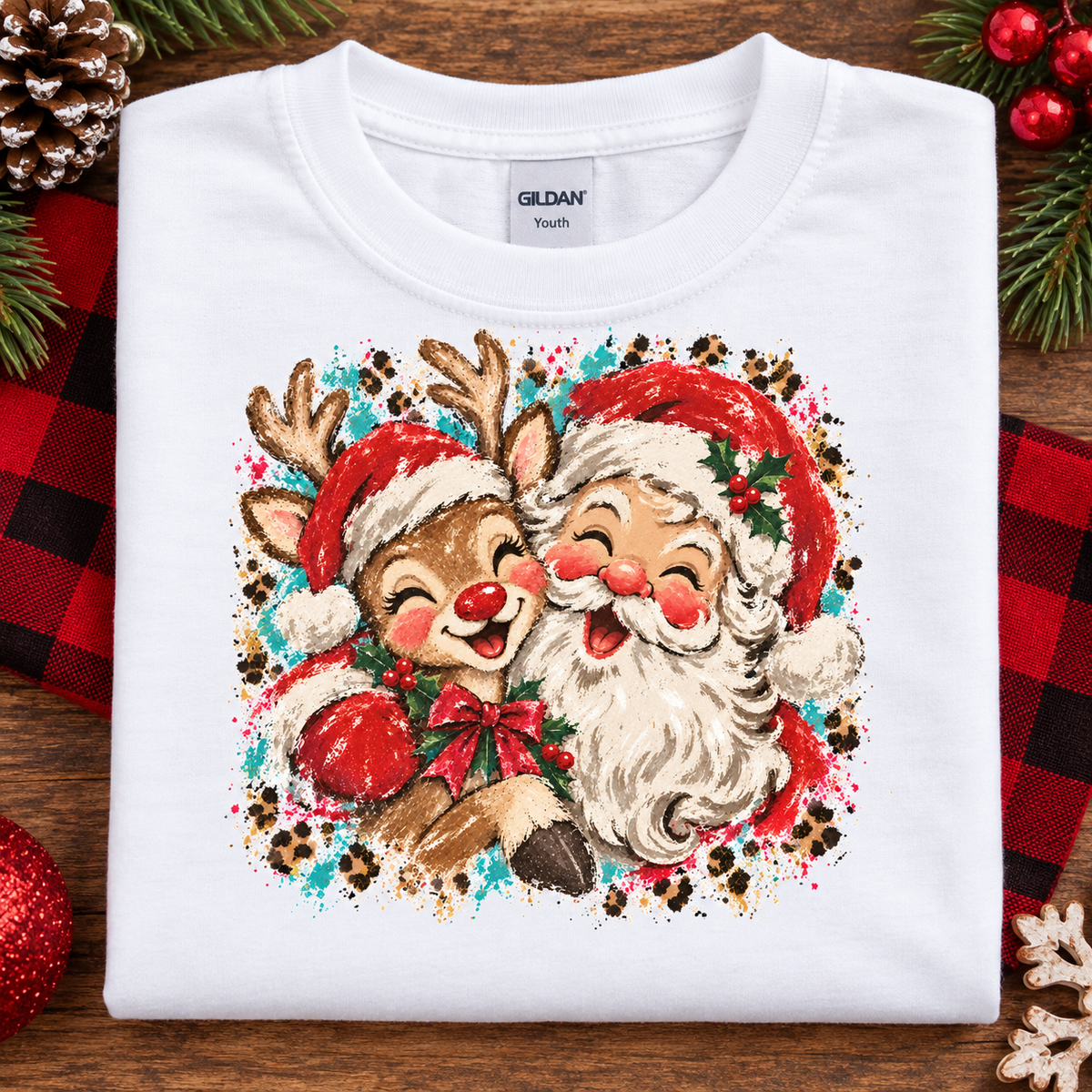 A folded white Gildan Youth t-shirt featuring a colorful graphic of a reindeer and Santa Claus hugging, styled on a rustic wooden background with holiday decorations.