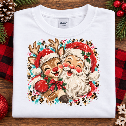 A folded white Gildan Youth t-shirt featuring a colorful graphic of a reindeer and Santa Claus hugging, styled on a rustic wooden background with holiday decorations.