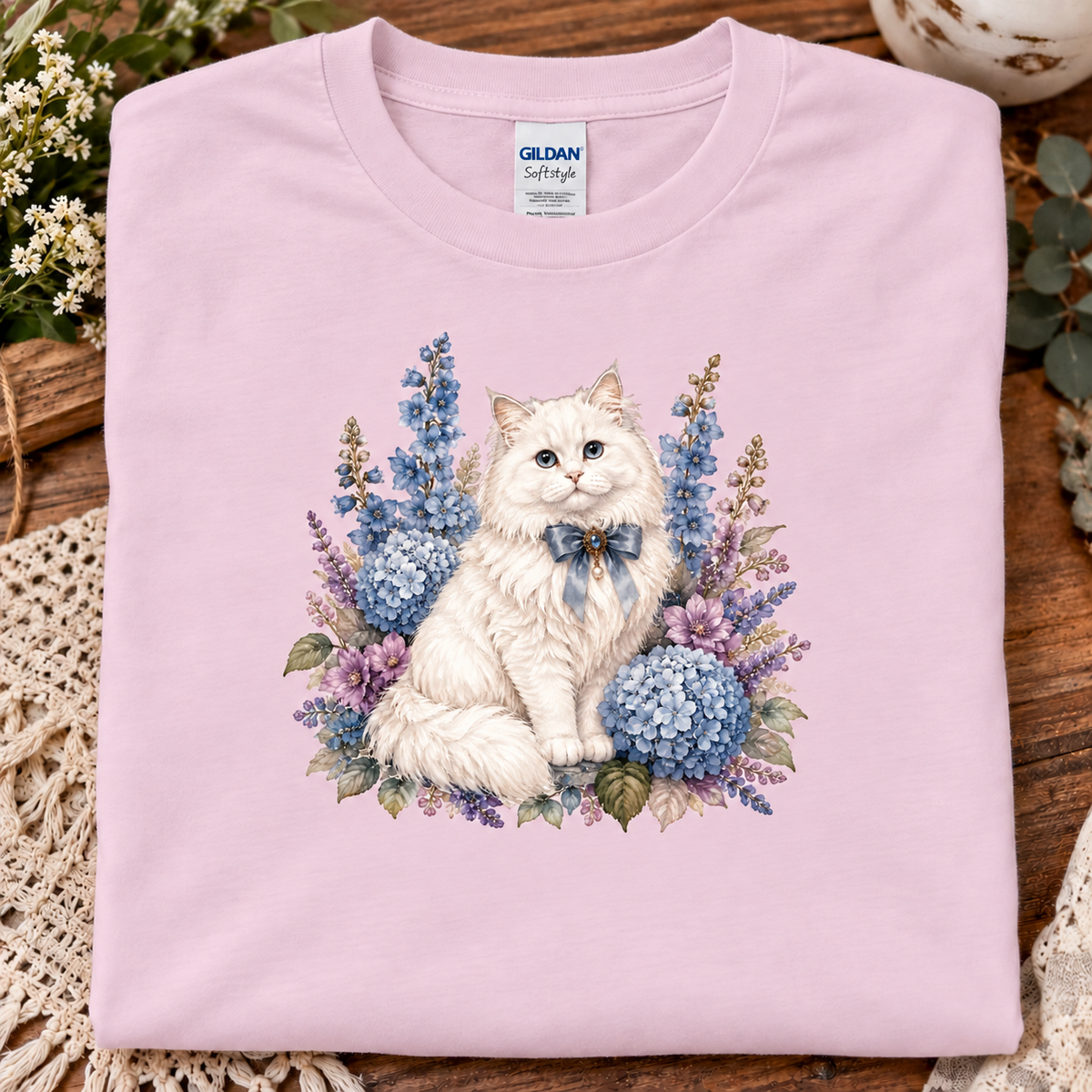 A folded light pink Gildan Softstyle t-shirt with a detailed graphic of a fluffy white cat surrounded by blue and purple hydrangeas, displayed on a rustic wooden surface with decorative flowers.