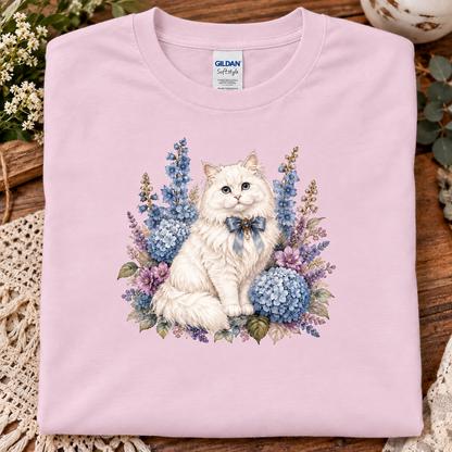 A folded light pink Gildan Softstyle t-shirt with a detailed graphic of a fluffy white cat surrounded by blue and purple hydrangeas, displayed on a rustic wooden surface with decorative flowers.