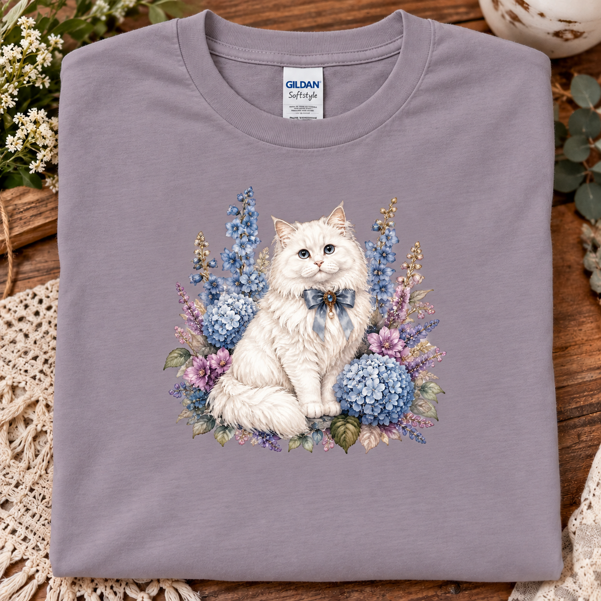 A folded Gildan Softstyle t-shirt in a paragon color, featuring a detailed graphic print of a fluffy white cat surrounded by blue and purple hydrangeas and flowers. 