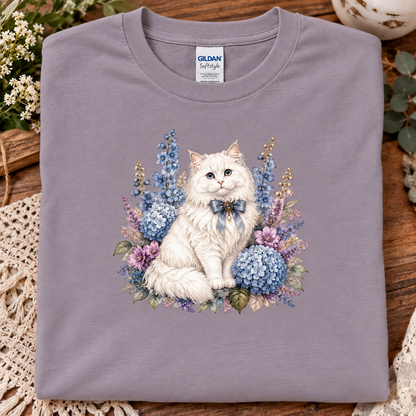 A folded Gildan Softstyle t-shirt in a paragon color, featuring a detailed graphic print of a fluffy white cat surrounded by blue and purple hydrangeas and flowers. 