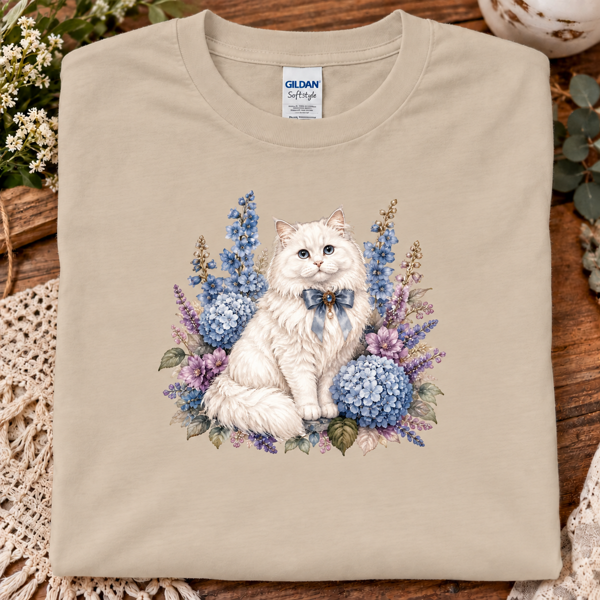 A sand Gildan Softstyle t-shirt featuring a watercolor-style graphic of a fluffy white cat wearing a blue bow, surrounded by blue and purple hydrangeas, laid flat on a rustic wooden surface with decorative lace and flowers.