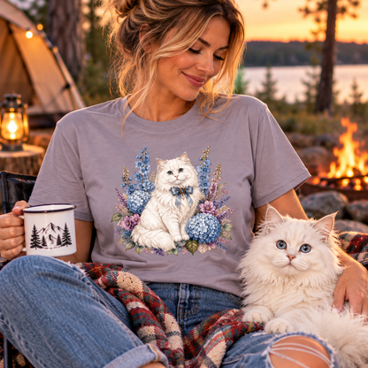 A woman sitting outdoors by a campfire at sunset wears a paragon colored graphic T-shirt featuring a white cat surrounded by blue and purple flowers. A fluffy white cat rests on her lap, and she holds a mug with a mountain design.