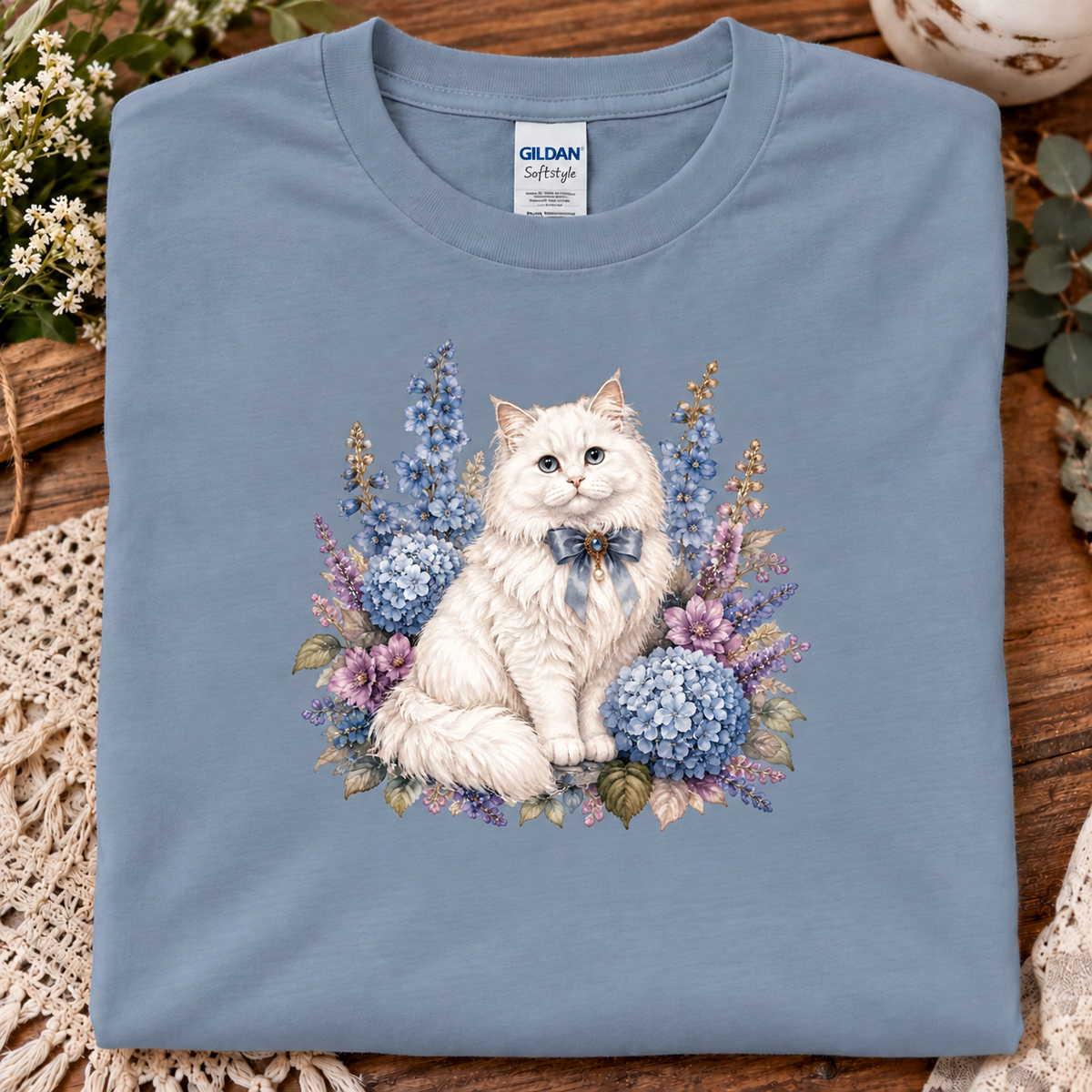 A folded stone blue Gildan Softstyle t-shirt featuring a detailed graphic of a fluffy white cat surrounded by blue and purple hydrangeas, displayed on a rustic wooden surface with decorative lace and flowers.