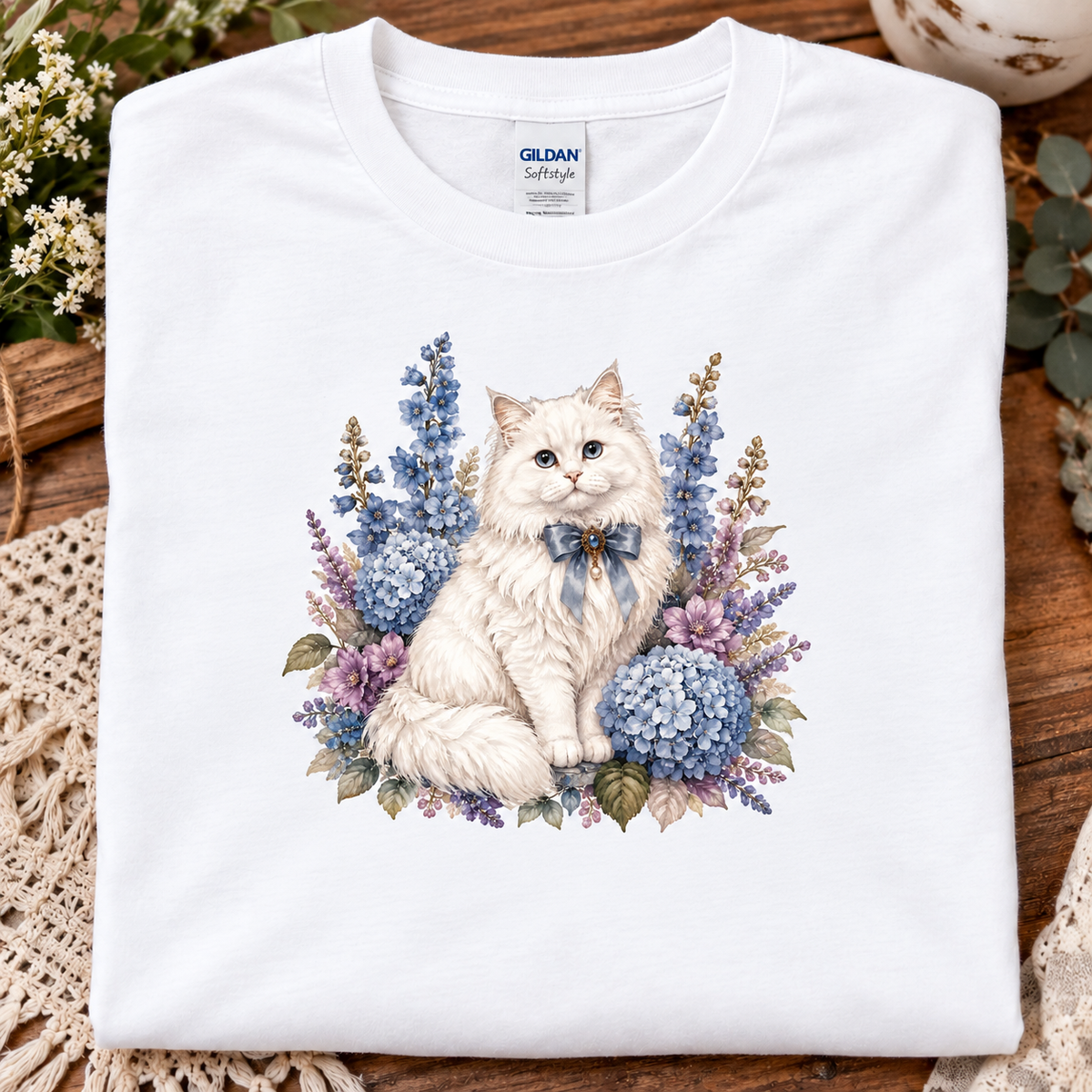 A folded white Gildan Softstyle T-shirt with a detailed graphic of a fluffy white cat surrounded by blue and purple flowers, displayed on a rustic wooden surface with decorative lace and flowers.