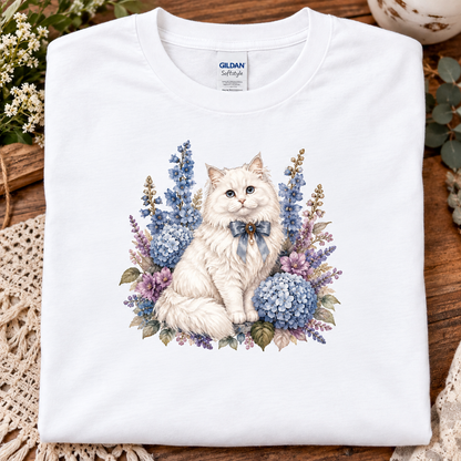 A folded white Gildan Softstyle T-shirt with a detailed graphic of a fluffy white cat surrounded by blue and purple flowers, displayed on a rustic wooden surface with decorative lace and flowers.