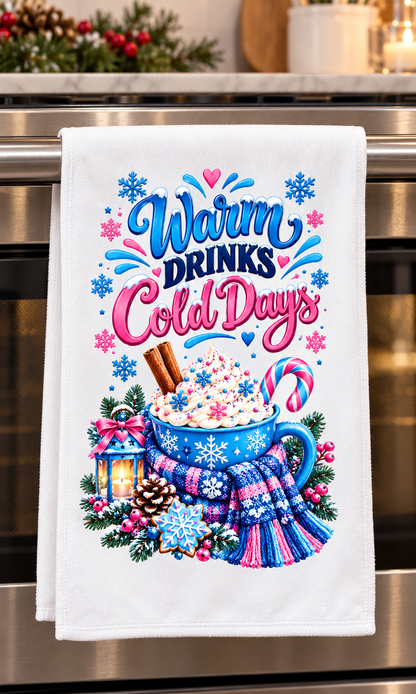 A white kitchen towel hanging on a stainless steel oven handle, featuring a colorful winter-themed graphic that reads 'Warm Drinks Cold Days' above a mug of hot cocoa with whipped cream, a scarf, snowflakes, and holiday decorations.