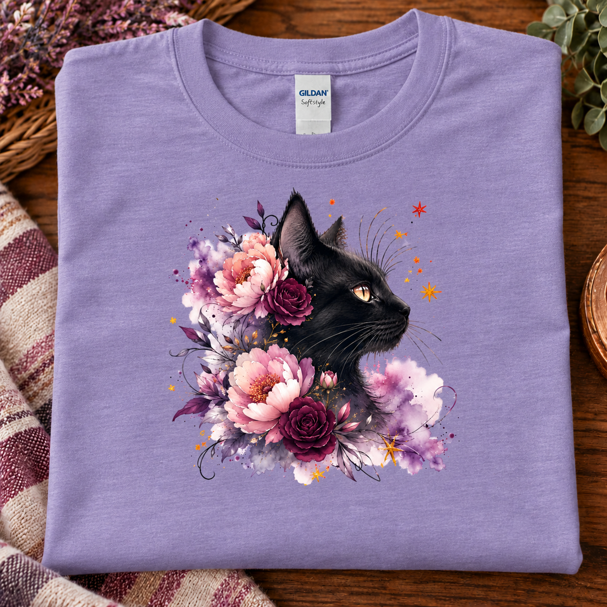 A folded heather purple t-shirt with a colorful graphic of a black cat surrounded by pink, purple, and white flowers, displayed on a wooden surface with a striped blanket and dried flowers.