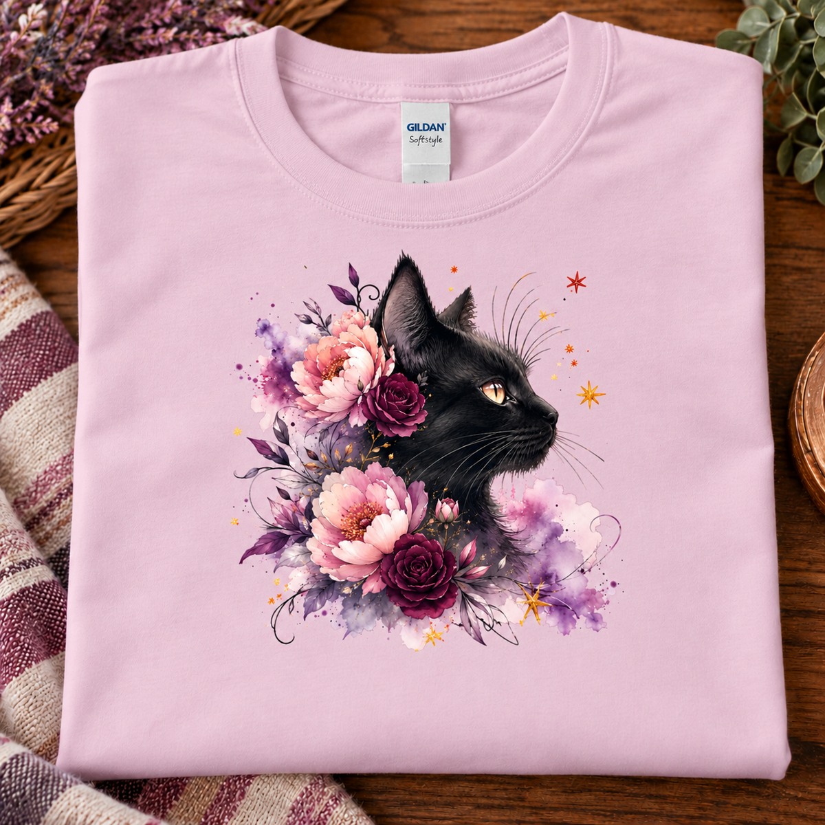 A folded light pink crewneck t-shirt with a graphic of a black cat surrounded by pink and purple flowers, displayed on a wooden surface with a striped blanket and dried flowers.
