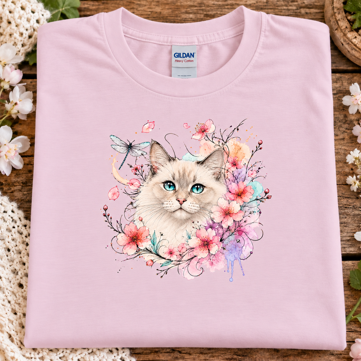 A folded light pink Gildan t-shirt with a colorful watercolor graphic of a cat surrounded by flowers and a dragonfly, displayed on a rustic wooden surface with decorative flowers.