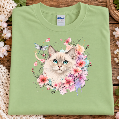 A folded pistachio green Gildan T-shirt with a colorful watercolor-style cat and floral print on the front, displayed on a rustic wooden surface with decorative flowers and lace around it.