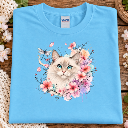 A folded sky blue Gildan t-shirt with a colorful graphic of a fluffy white cat surrounded by pink and purple flowers, displayed on a rustic wooden surface with decorative flowers.