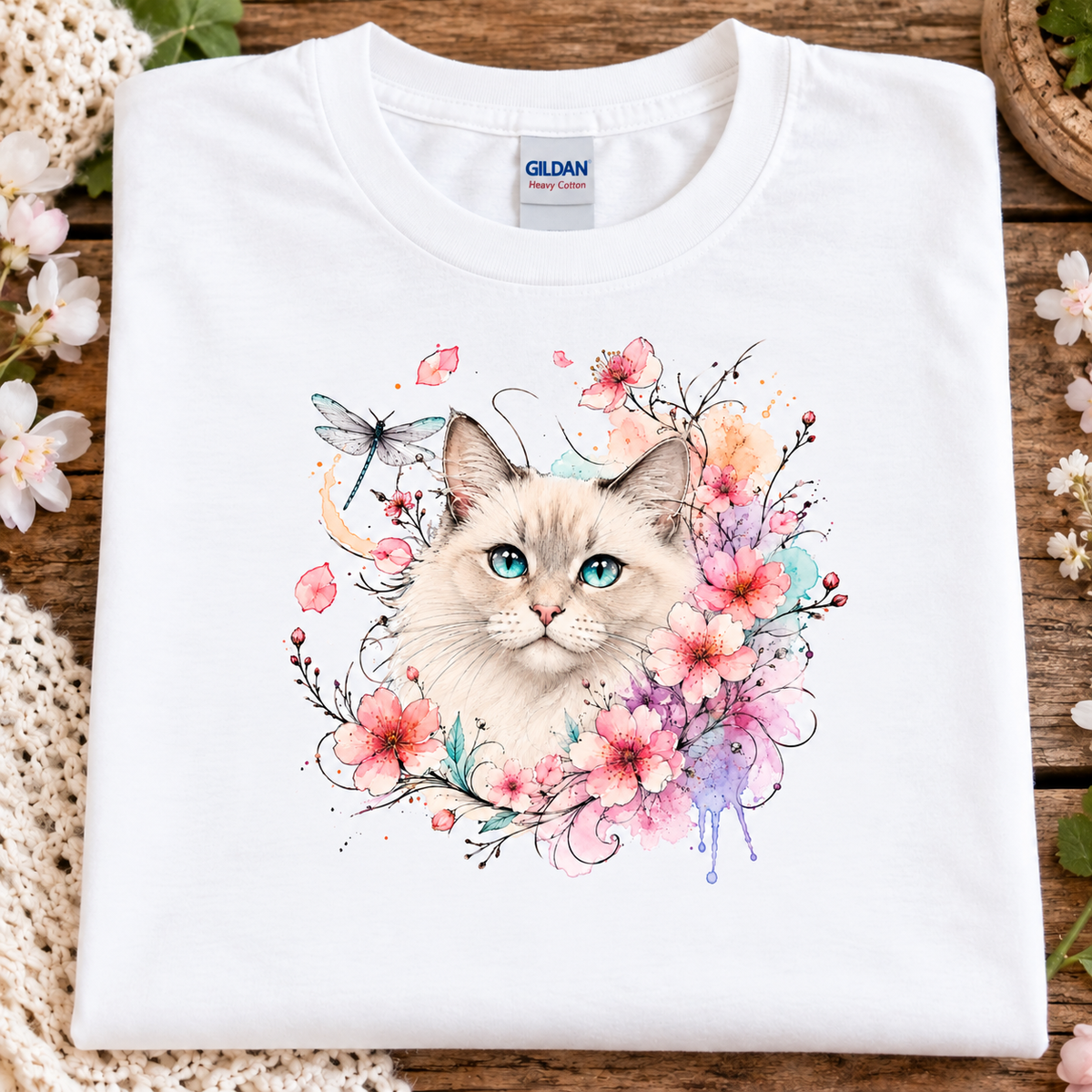 A folded white Gildan t-shirt with a colorful watercolor graphic of a fluffy cream-colored cat surrounded by pink and purple flowers, displayed on a rustic wooden surface with decorative flowers.