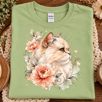 A folded pistachio green Gildan Softstyle t-shirt featuring a detailed watercolor-style graphic of a fluffy cream-colored cat surrounded by pink and peach flowers, displayed on a rustic wooden background with a crochet mat.