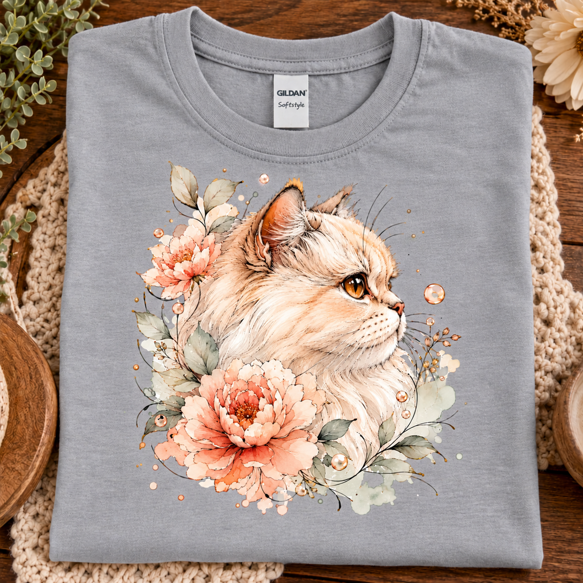 A folded sport gray Gildan Comfort T-shirt with a detailed watercolor-style print of a fluffy cream-colored cat surrounded by peach and pink flowers, green leaves, and small gold dots, displayed on a rustic wooden surface with woven placemats.