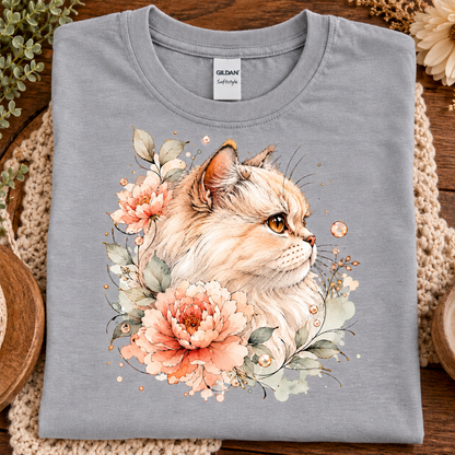 A folded sport gray Gildan Comfort T-shirt with a detailed watercolor-style print of a fluffy cream-colored cat surrounded by peach and pink flowers, green leaves, and small gold dots, displayed on a rustic wooden surface with woven placemats.