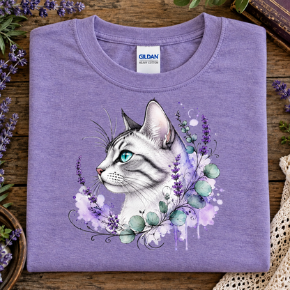 A folded heather purple Gildan cotton t-shirt with a graphic of a white tabby cat surrounded by purple flowers and green leaves, displayed on a rustic wooden surface with decorative flowers and a lace cloth.