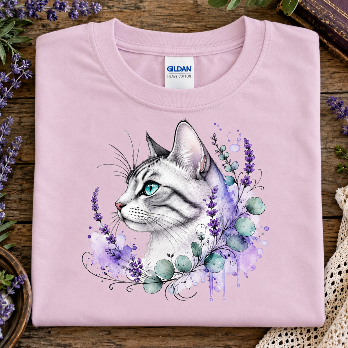 A folded light pink Gildan cotton t-shirt with a detailed graphic of a white and gray tabby cat with blue eyes, surrounded by purple lavender and green leaves, displayed on a rustic wooden surface with decorative flowers.