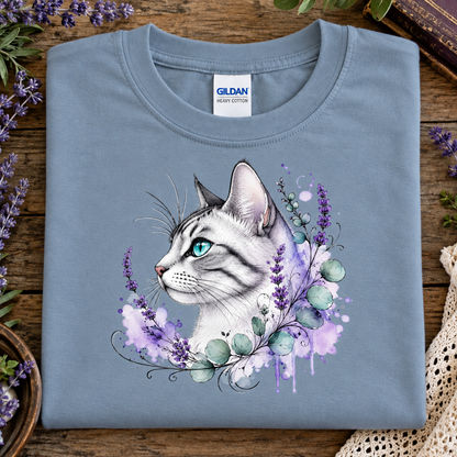 A folded stone blue Gildan cotton t-shirt with a watercolor-style graphic of a white cat with green eyes surrounded by purple lavender and green leaves, displayed on a rustic wooden surface with decorative flowers.