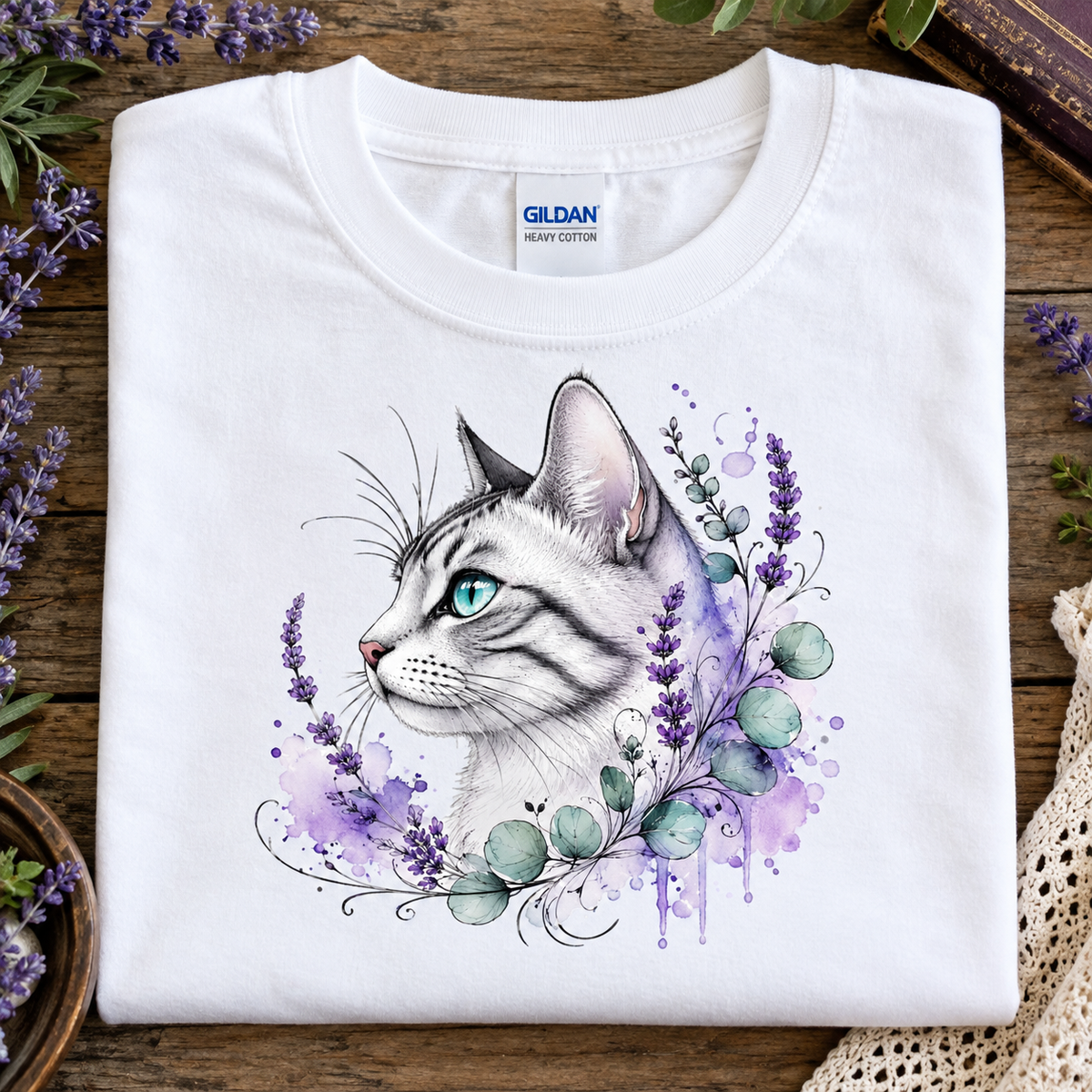 A folded white Gildan Comfort Colors t-shirt with a detailed graphic of a white cat with blue eyes surrounded by purple flowers and green leaves, displayed on a rustic wooden surface with decorative lavender and lace.