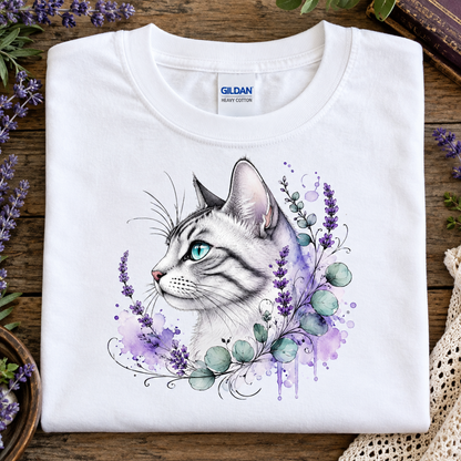A folded white Gildan Comfort Colors t-shirt with a detailed graphic of a white cat with blue eyes surrounded by purple flowers and green leaves, displayed on a rustic wooden surface with decorative lavender and lace.