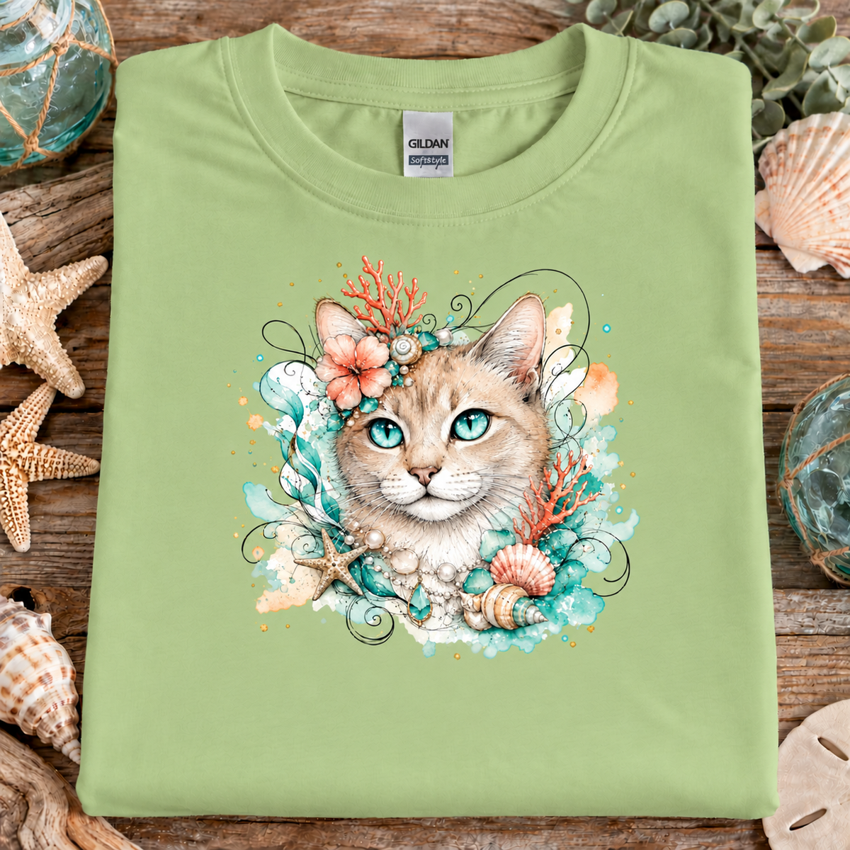 A pistachio green colored t-shirt with a tan tabby cat surrounded by seashells