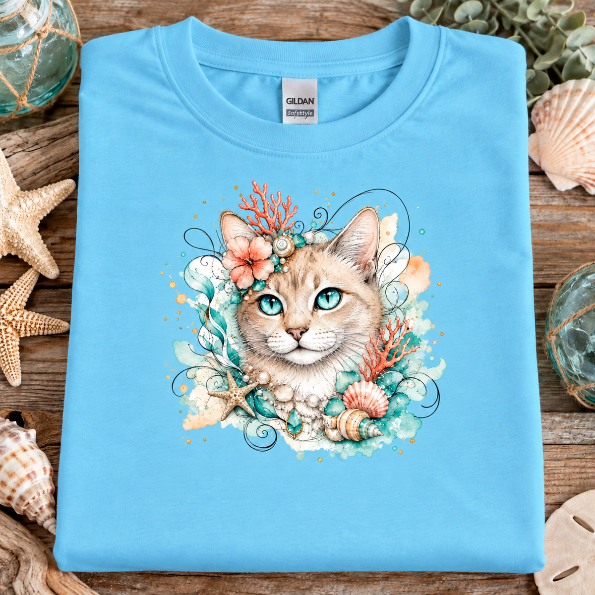 A sky blue colored t-shirt with a tan tabby cat surrounded by seashells