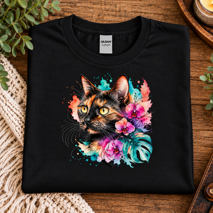 A black t-shirt with a watercolor-style graphic design of a calico cat head surrounded by pink orchids, green monstera leaves, and colorful paint splashes on a white background.