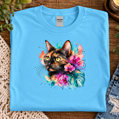 A sky blue t-shirt with a watercolor-style graphic design of a calico cat head surrounded by pink orchids, green monstera leaves, and colorful paint splashes on a white background