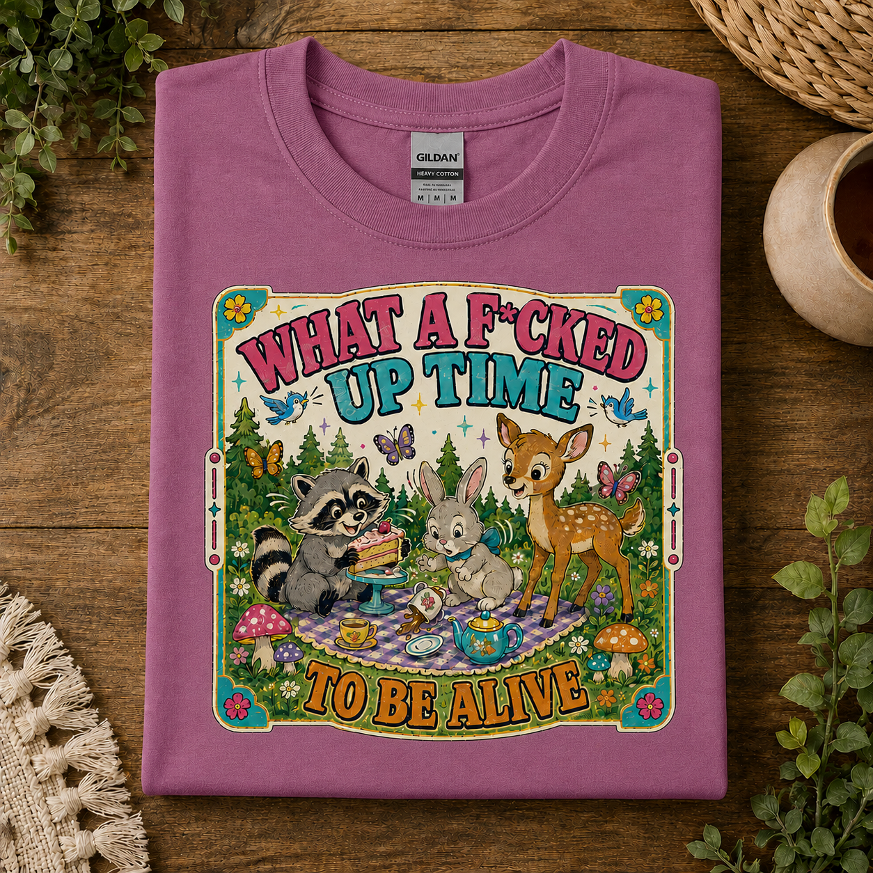 A folded heather radiant orchid colored  Gildan t-shirt with a colorful graphic print of woodland animals having a picnic, displayed on a rustic wooden background with decorative plants and a woven basket.