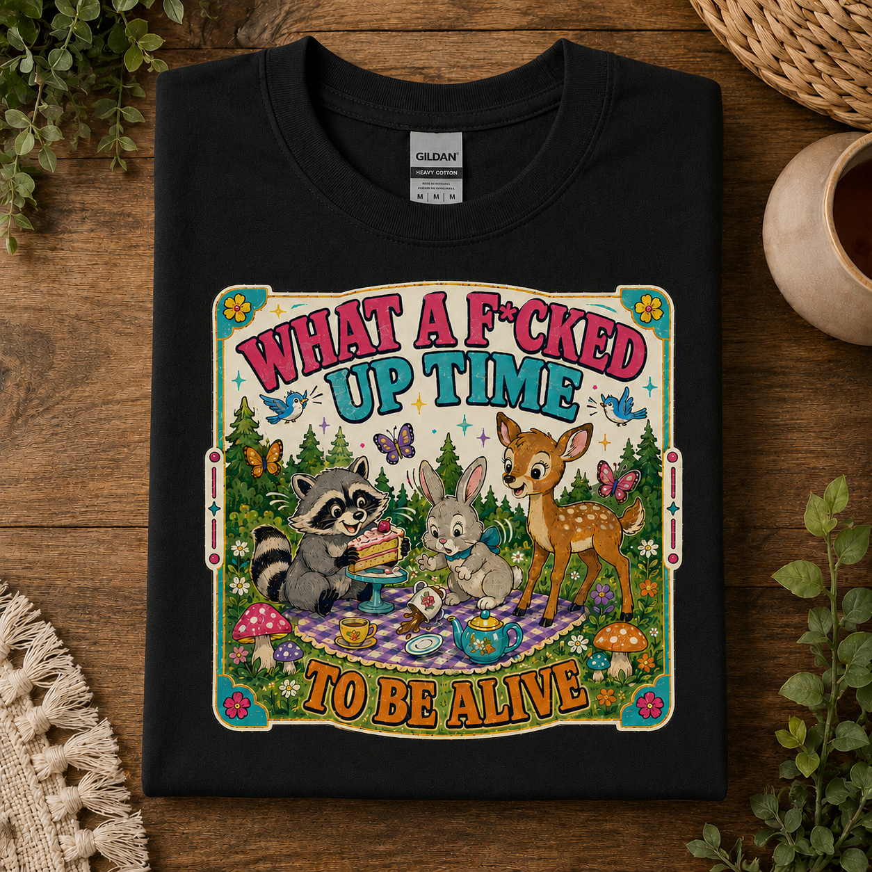 A black Gildan t-shirt laid flat on a rustic wooden surface, featuring a colorful cartoon graphic of a raccoon, rabbit, and deer having a picnic with the text 'WHAT A F*CKED UP TIME TO BE ALIVE'.